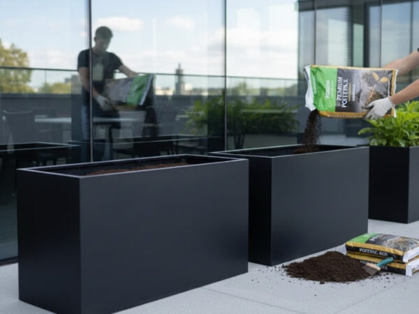 Person pouring potting mix into a large black planter on a modern balcony.